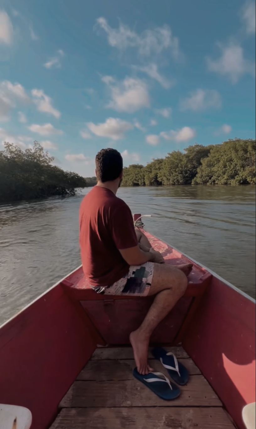 Ricardo Longo de costas em um barco no rio, atravessando paisagem com manguezal — entre a estrada e o sistema